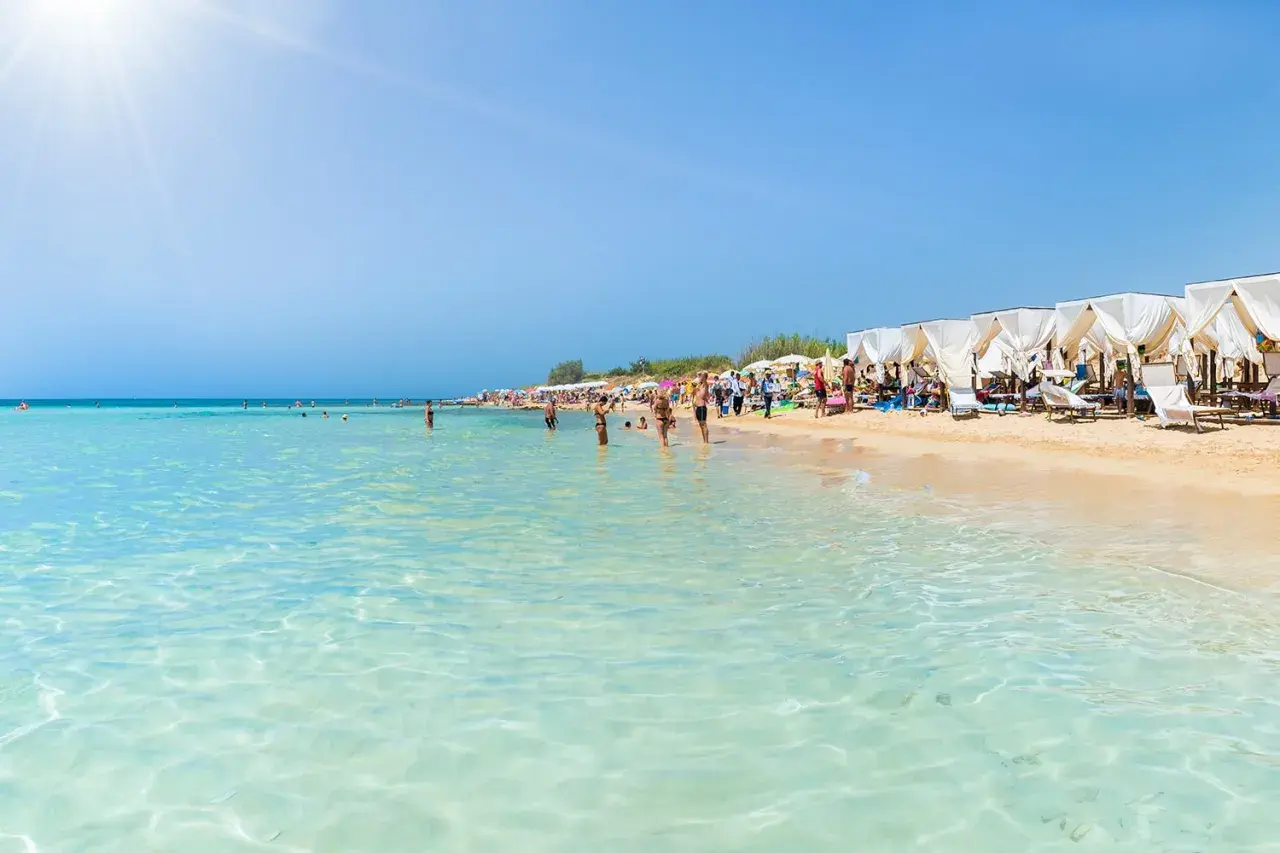 Spiaggia affollata in Puglia, con gente che fa il bagno nelle acque cristalline e si rilassa sotto i gazebo bianchi.