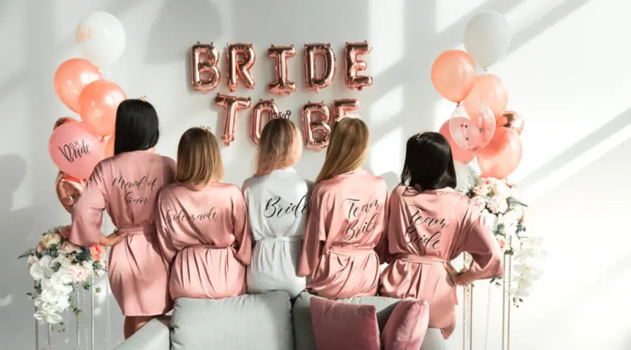 Pięć kobiet w satynowych szatkach, idealny prezent na wieczór panieński z klasą. Balony i napisy "Bride to Be" tworzą świąteczną atmosferę.
