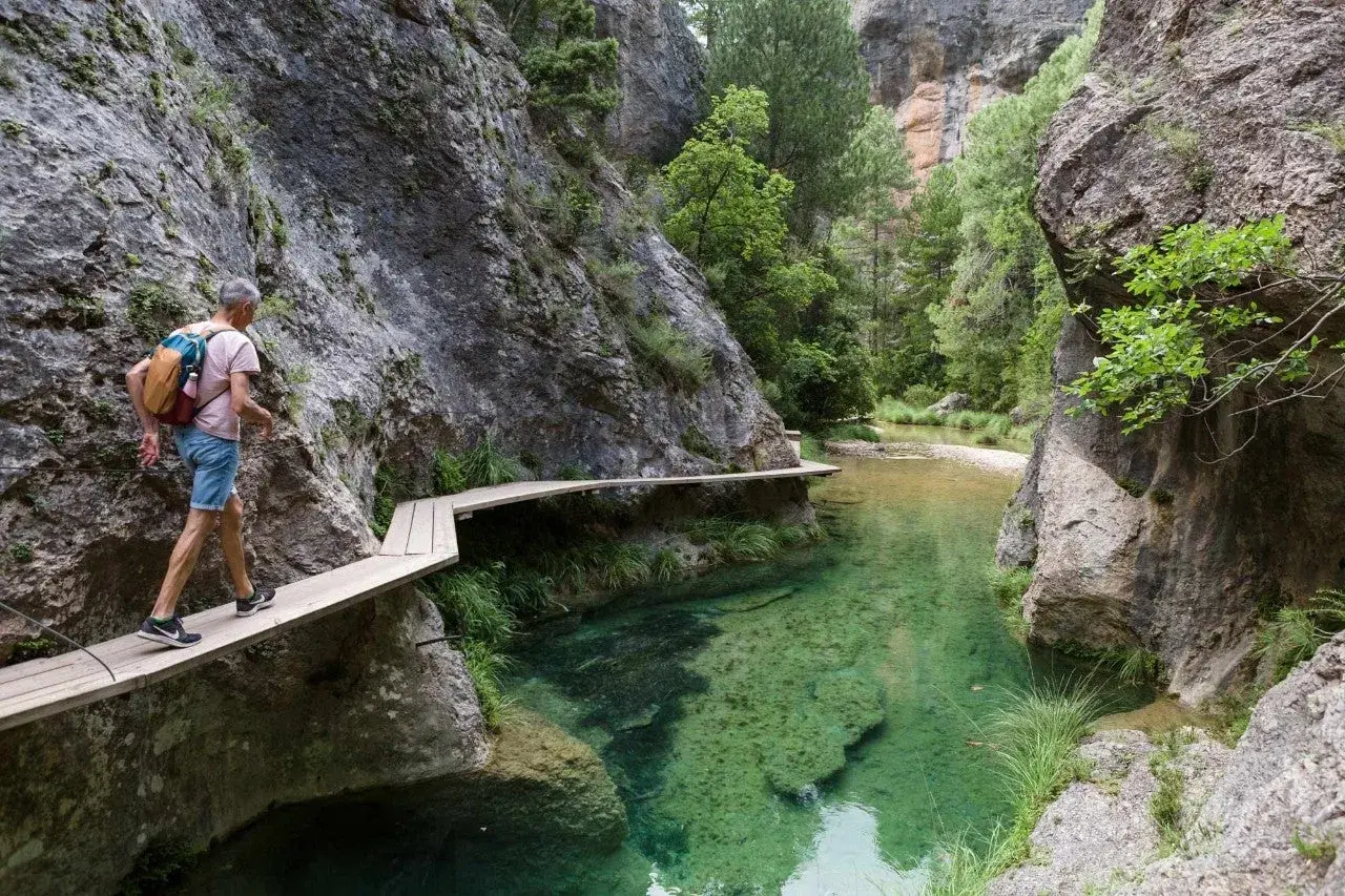 Hombre camina por pasarela de madera en la ruta parrizal de beceite, junto a un río de aguas cristalinas y paredes rocosas.