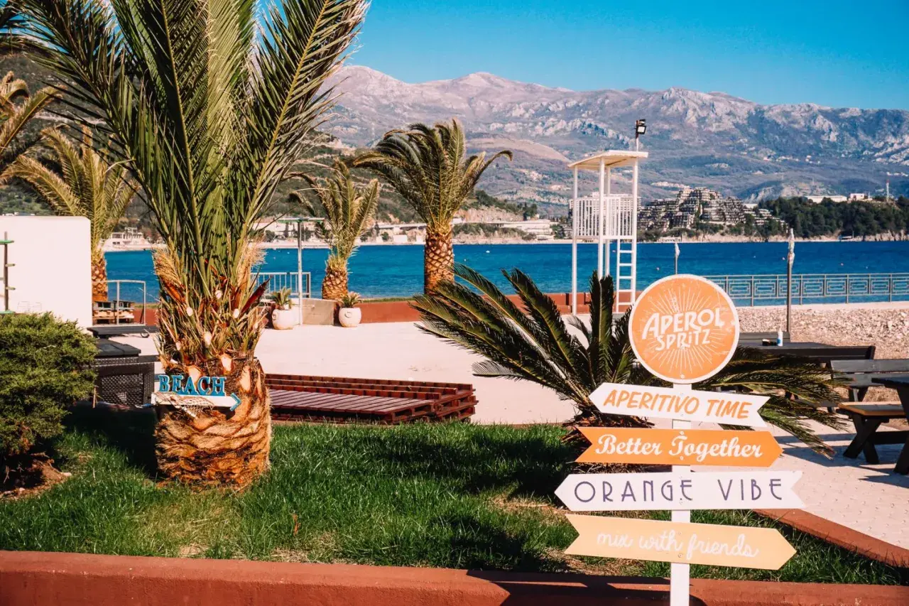 Palmy, morze i góry w Czarnogórze. Znaki wskazują plażę, Aperol Spritz i "Orange Vibe". Idealne miejsce na wakacje.