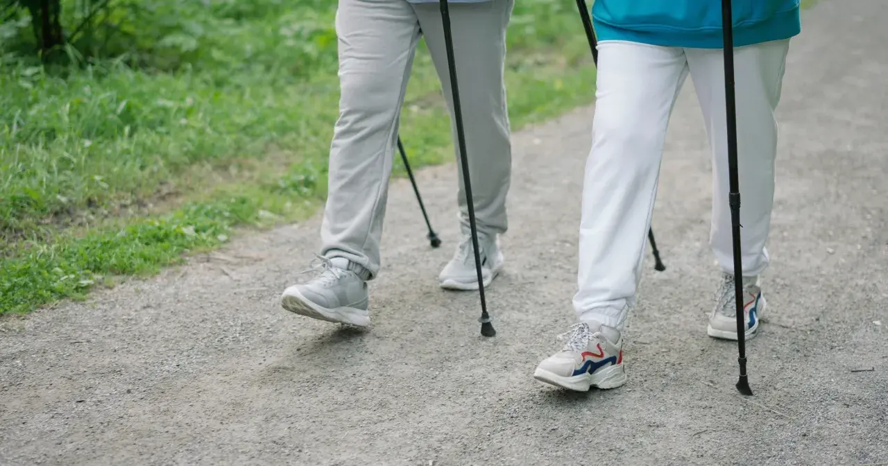 Dwie osoby uprawiające **nordic walking** na żwirowej ścieżce, otoczone zielenią.