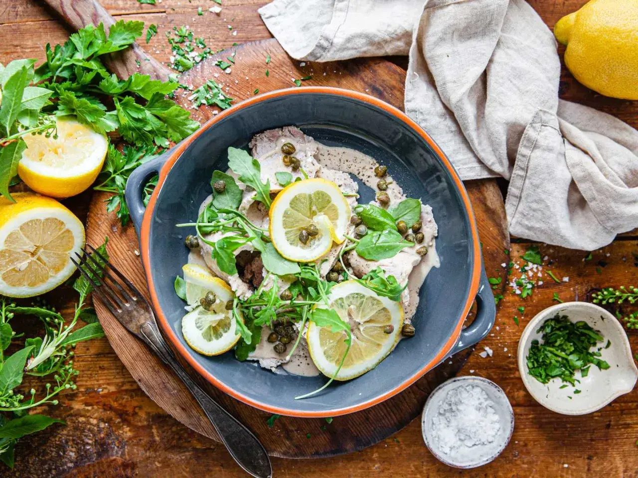 Vitello tonnato i en grå skål, garnerad med citronskivor, kapris och ruccola. Serveras med färska örter och citroner på ett träbord.