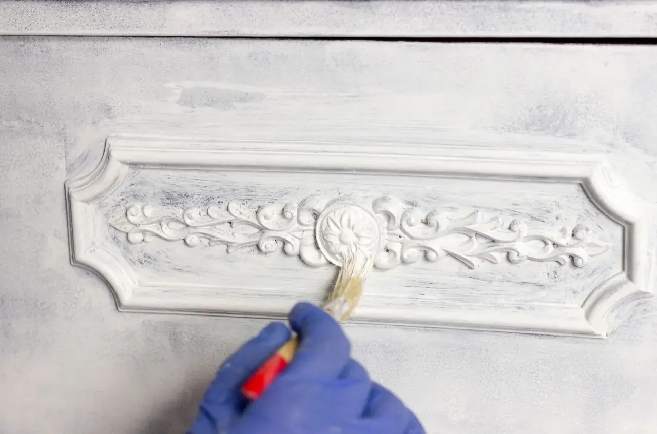 Mano con guanto blu che applica chalk paint fai da te su un mobile con decorazioni in rilievo, creando un effetto shabby chic.