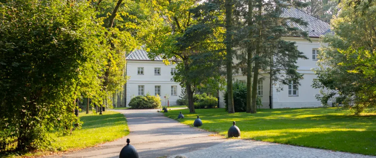 Biały dworek otoczony zielenią, z alejką i ozdobnymi kulami. Miejsce, gdzie nagrywano "Nad Niemnem".