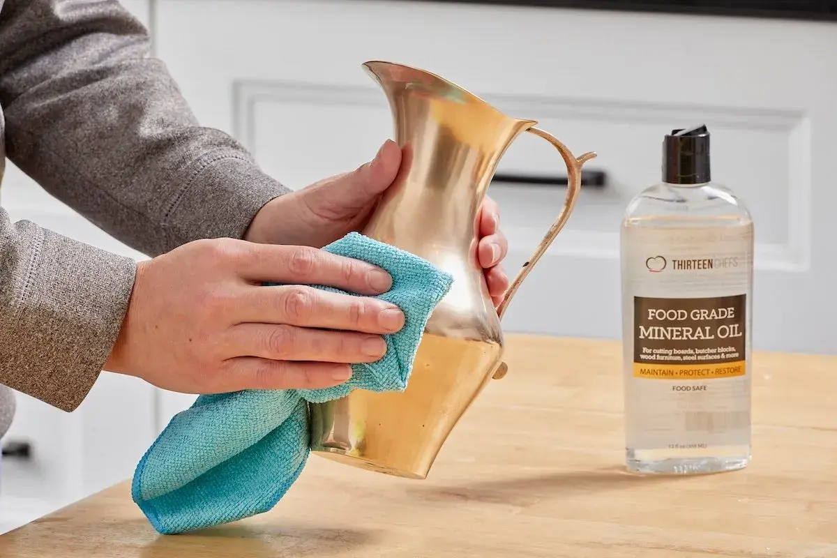 Hands polish a brass pitcher with a blue cloth, showing how to clean brass with mineral oil.