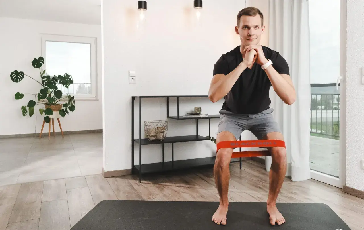Mann macht Kniebeugen mit Widerstandsband. Die tiefe Flexion im Knie wird durch das Band unterstützt und erschwert.