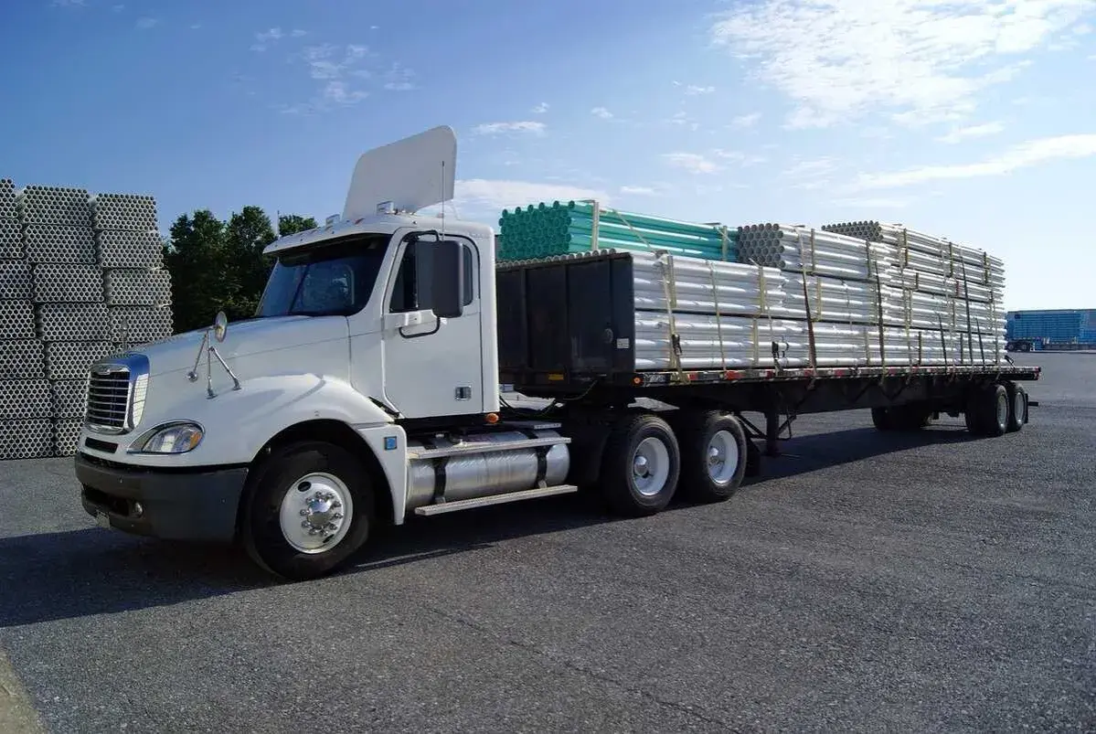 Camion blanc prêt pour le transport, avec un chargement de tuyaux blancs et verts bien sécurisé. L'arrimage des charges est essentiel pour un trajet sûr.