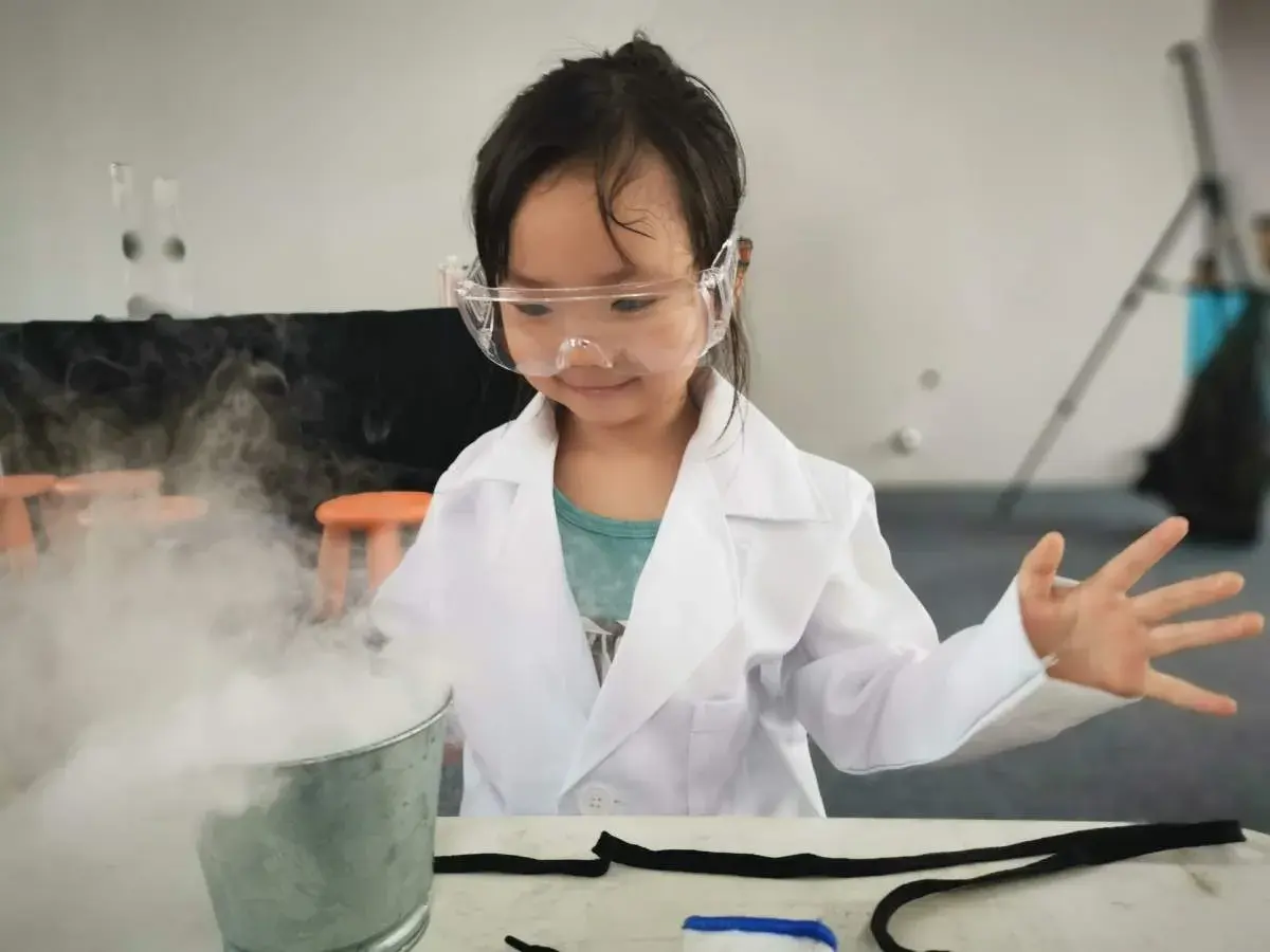 Niña con bata y gafas realiza experimentos para niños de 7 años, creando humo blanco de un cubo.