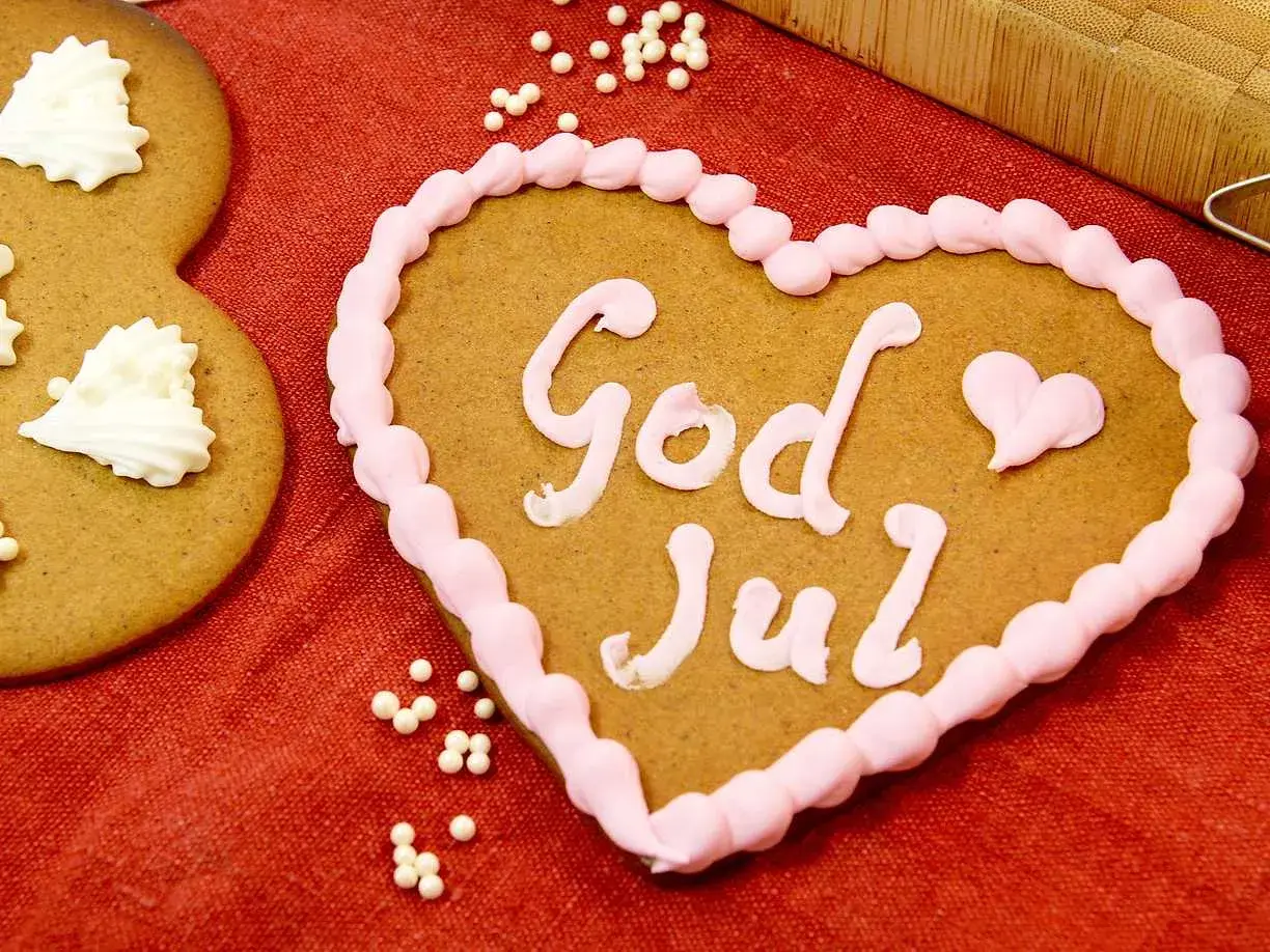 Hjärtformade pepparkakor med rosa glasyr och texten "God Jul". Bredvid syns en pepparkaka dekorerad med vita pepparkakor-liknande mönster.