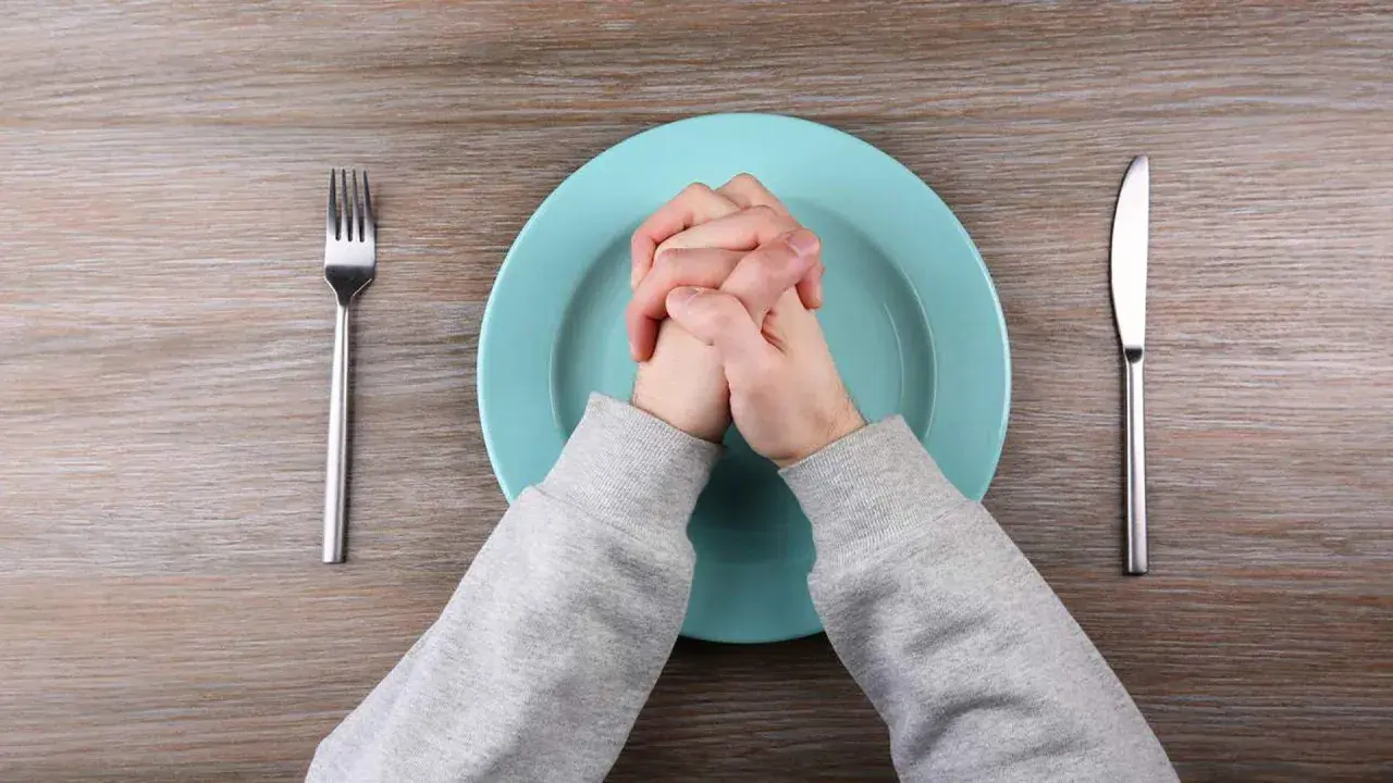 Manos entrelazadas sobre plato vacío, tenedor y cuchillo a los lados. Quizás piensa que no cenar adelgaza.