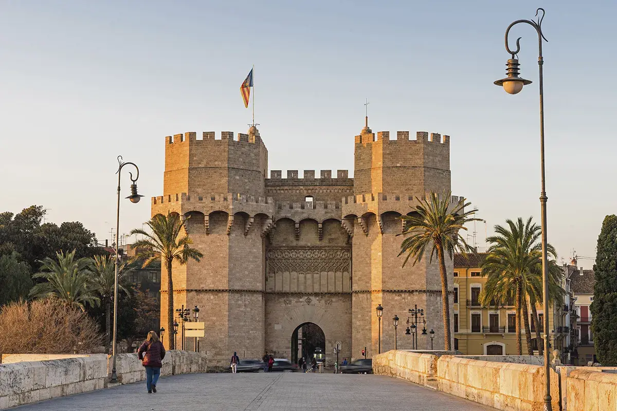 Las **Torres de Serranos**, imponentes puertas de la antigua muralla de Valencia, se alzan majestuosas bajo un cielo despejado. Palmeras y farolas flanquean el camino.