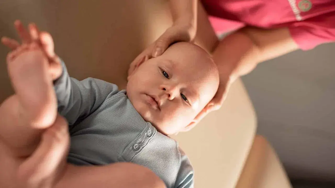 Niemowlę w niebieskim body, któremu fizjoterapeuta delikatnie masuje główkę.