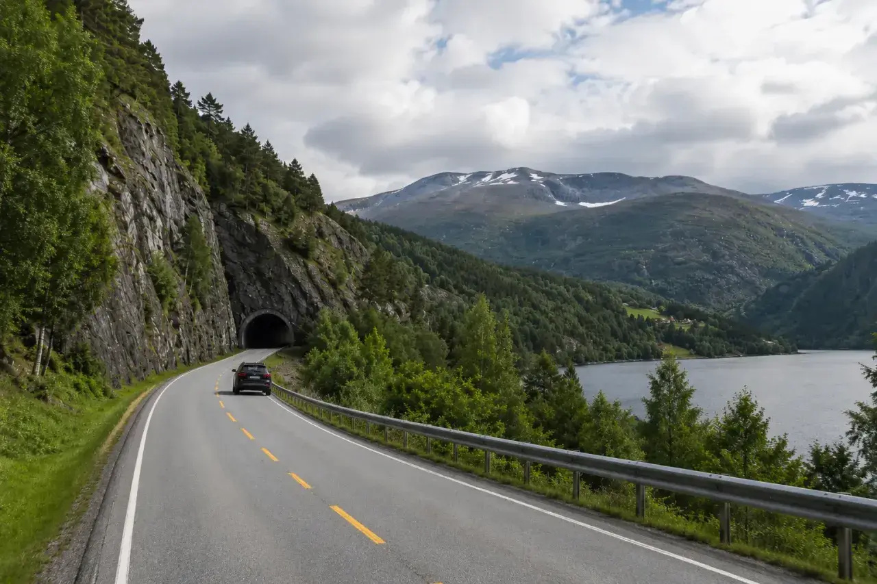 Svensk bil på väg mot en tunnel i Norge. Vacker bergig natur med sjö och moln.