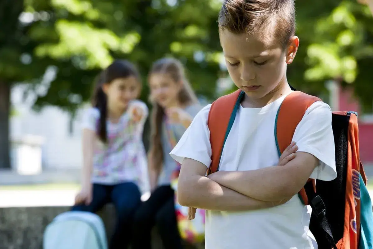 Ein Junge mit Rucksack steht traurig da, während zwei Mädchen ihn im Hintergrund auslachen. Ein trauriges Bild von Mobbing in der Grundschule.