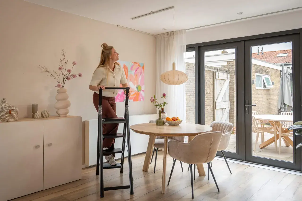 Mujer en escalera ajustando una lámpara colgante de dos luces sobre una mesa de comedor.