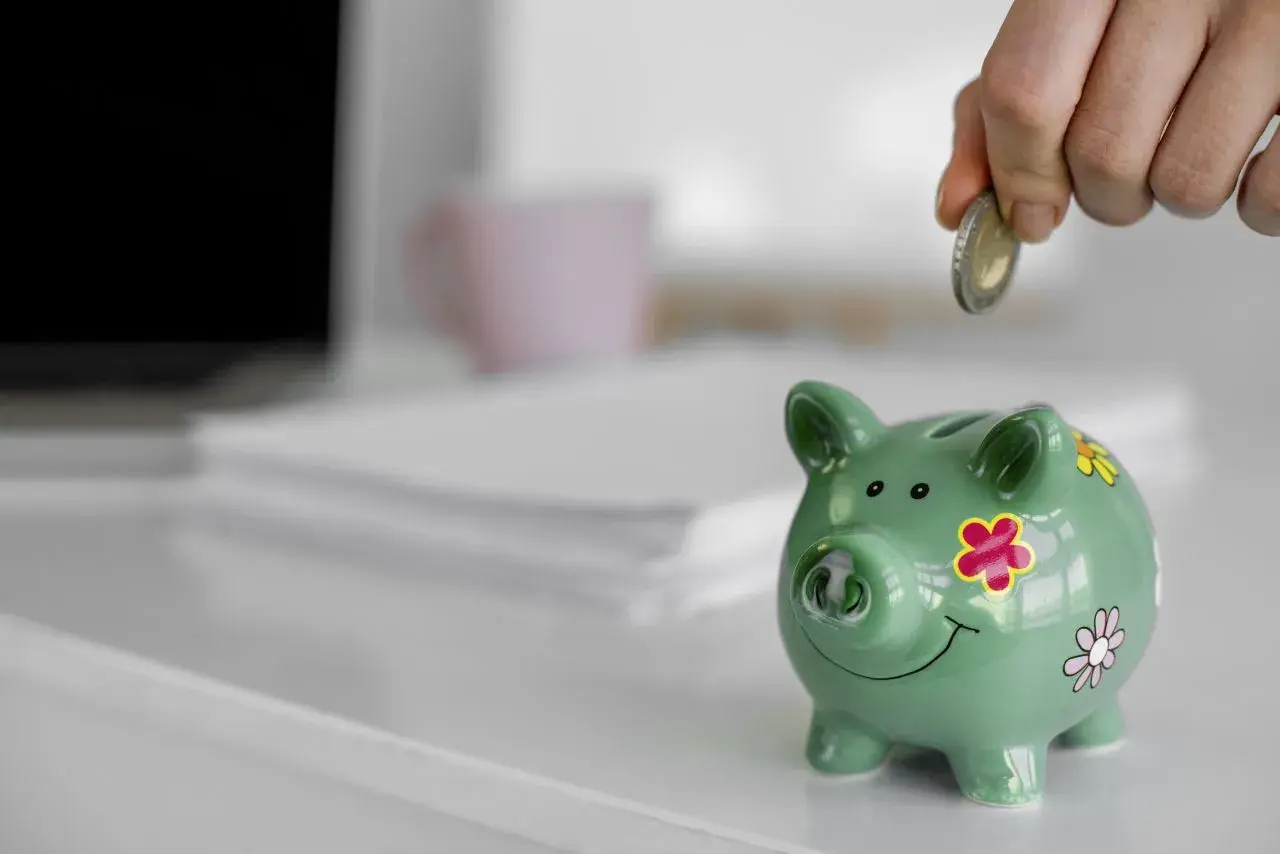 Ręka wrzuca monetę do zielonej świnki skarbonki. To symbol oszczędzania i finansowego zabezpieczenia.