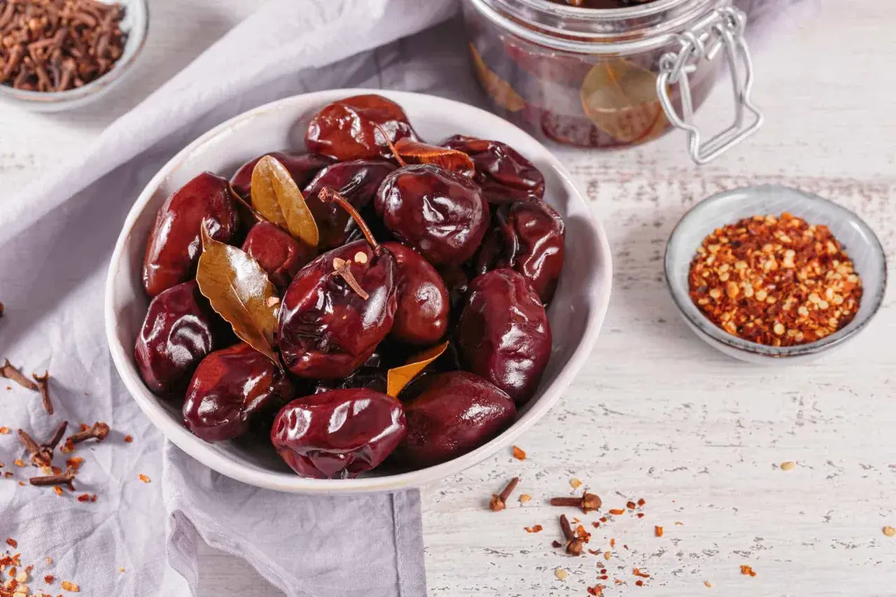 Schwarze, glänzende, fermentierte Pflaumen in einer Schüssel, bereit zum Genießen. Gewürze wie Nelken und Chiliflocken deuten darauf hin, wie man fermentierte Pflaumen selber machen kann.