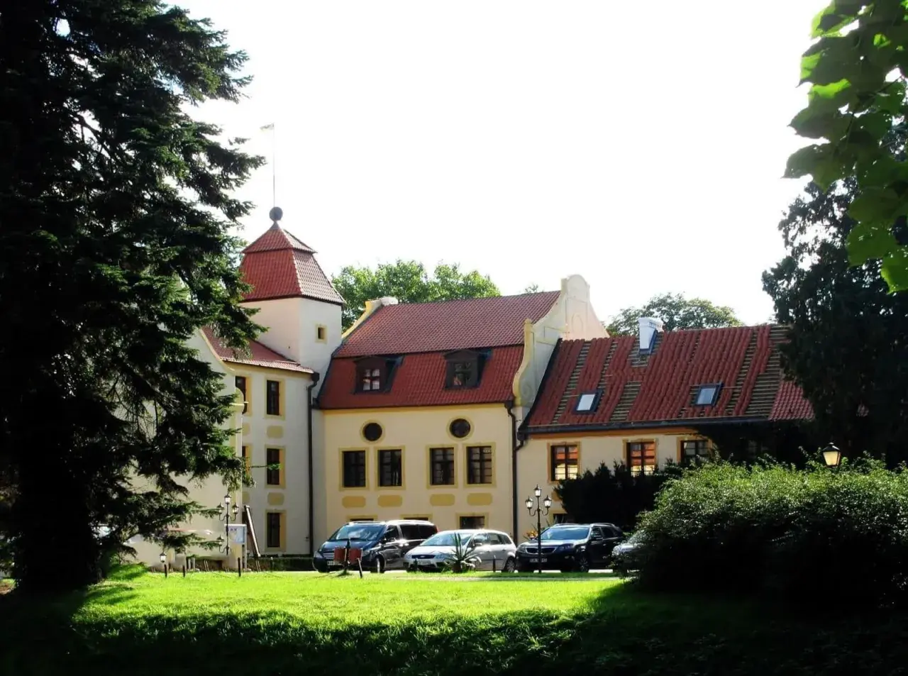 Pałac w Krokowej z czerwoną wieżą i dachami, otoczony zielenią. Przed budynkiem zaparkowane samochody.