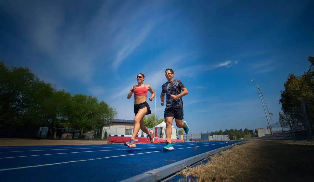 Coppia corre su pista blu, un allenamento intenso per la loro prossima corsa a tappe. Cielo azzurro e nuvole bianche.