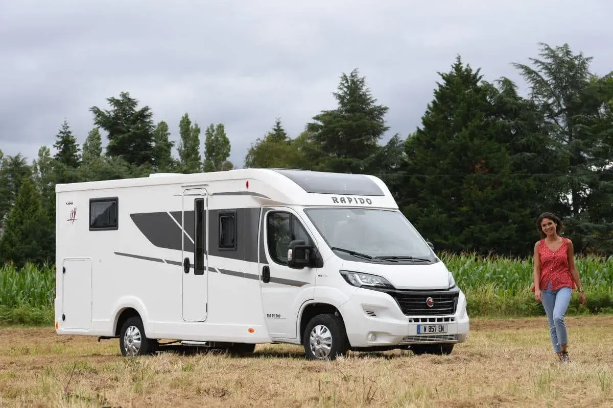 Un camping-car Rapido C86 blanc, moderne, garé dans un champ. Une jeune femme souriante se tient à droite. Avis Rapido C86.