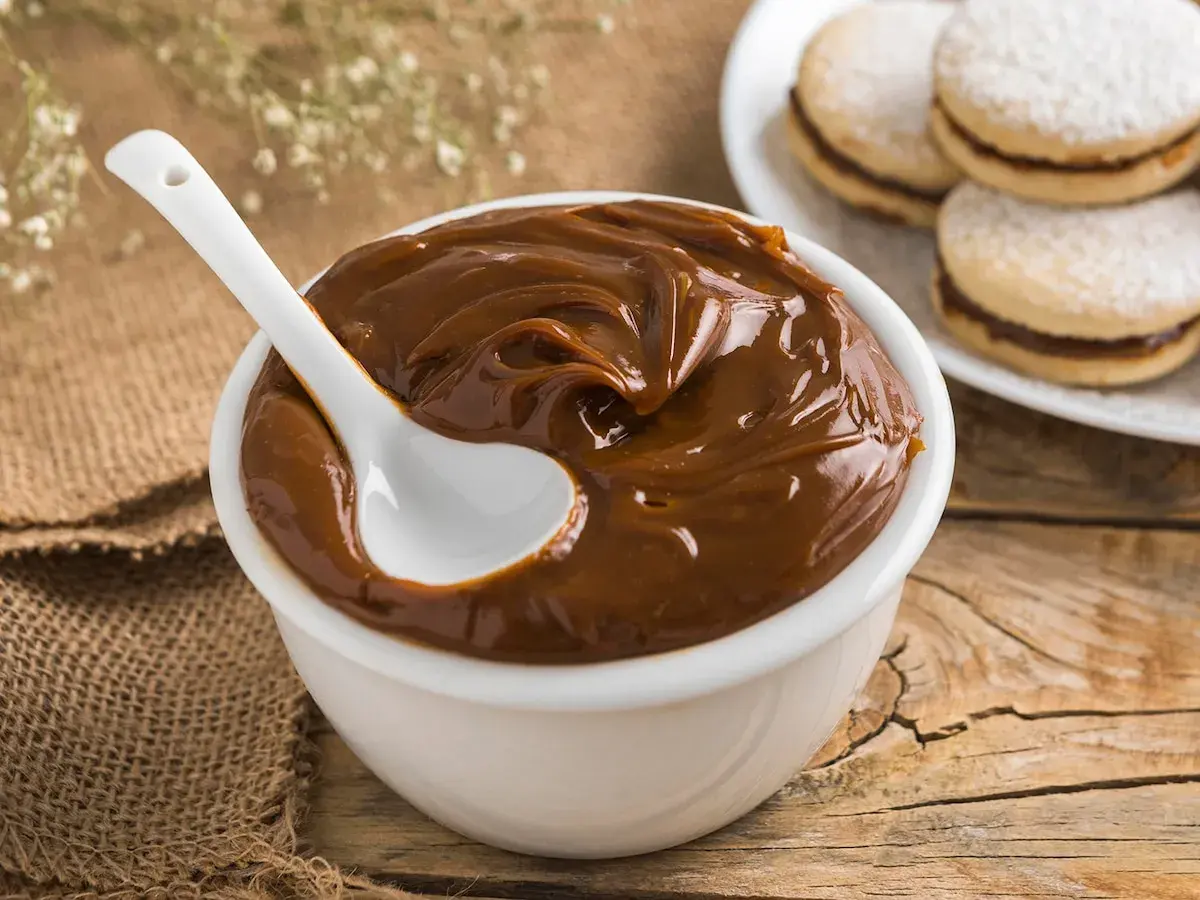 Un tazón blanco lleno de dulce de leche cremoso, con una cuchara blanca. Al fondo, alfajores espolvoreados con azúcar glas.