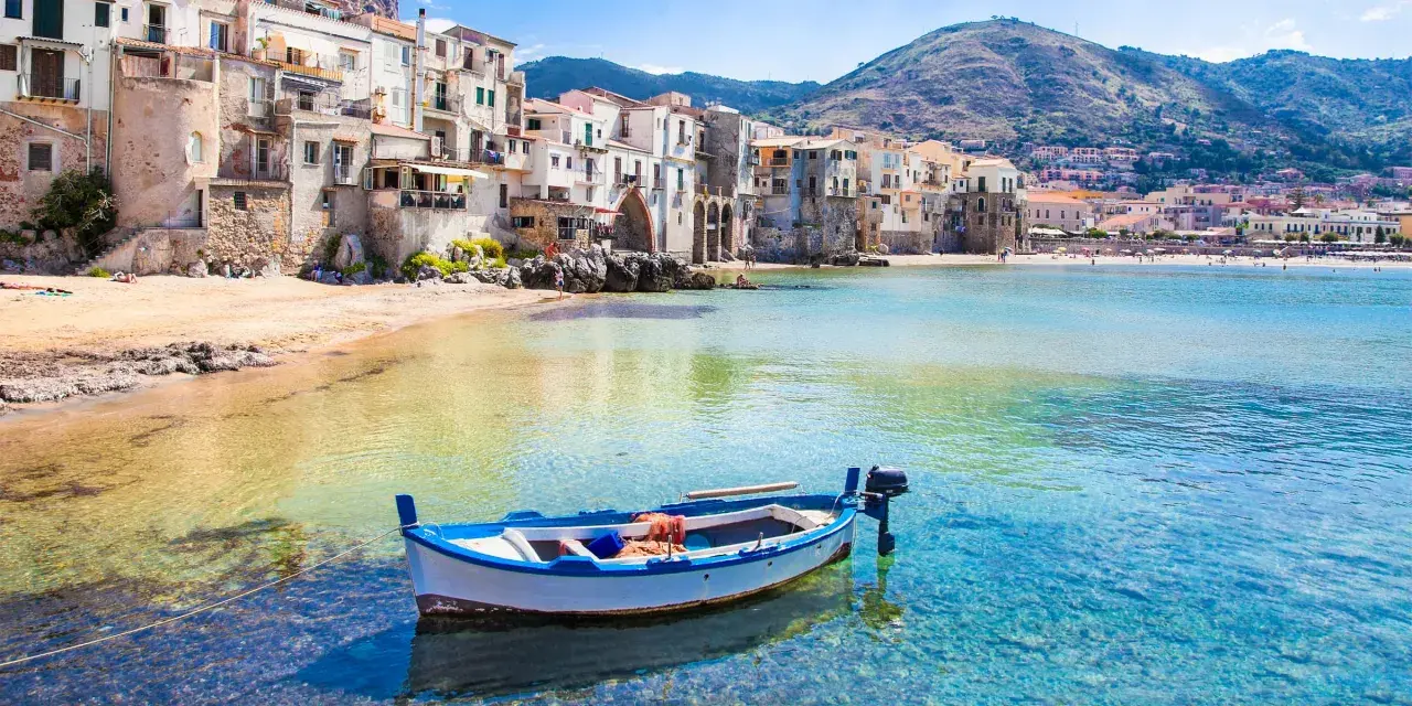Bateau blanc et bleu ancré dans les eaux cristallines de la plage de Cefalù, avec des maisons colorées en arrière-plan.