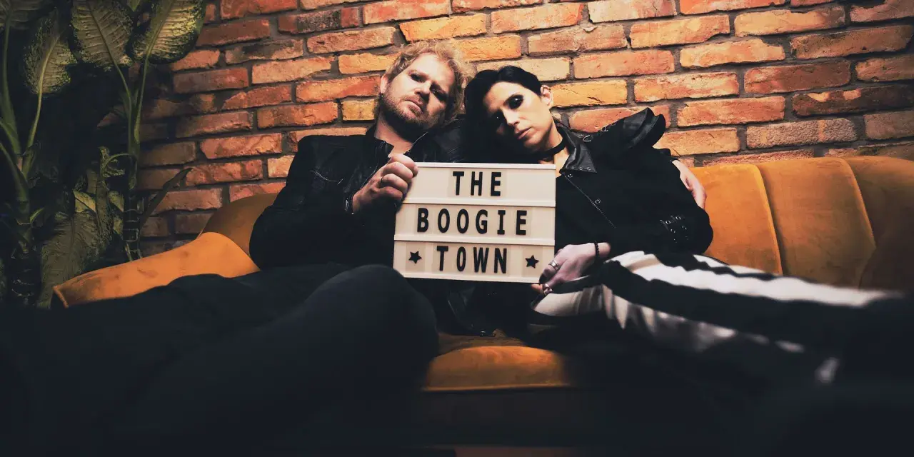 Para siedzi na kanapie, trzymając tabliczkę z napisem "THE BOOGIE TOWN".