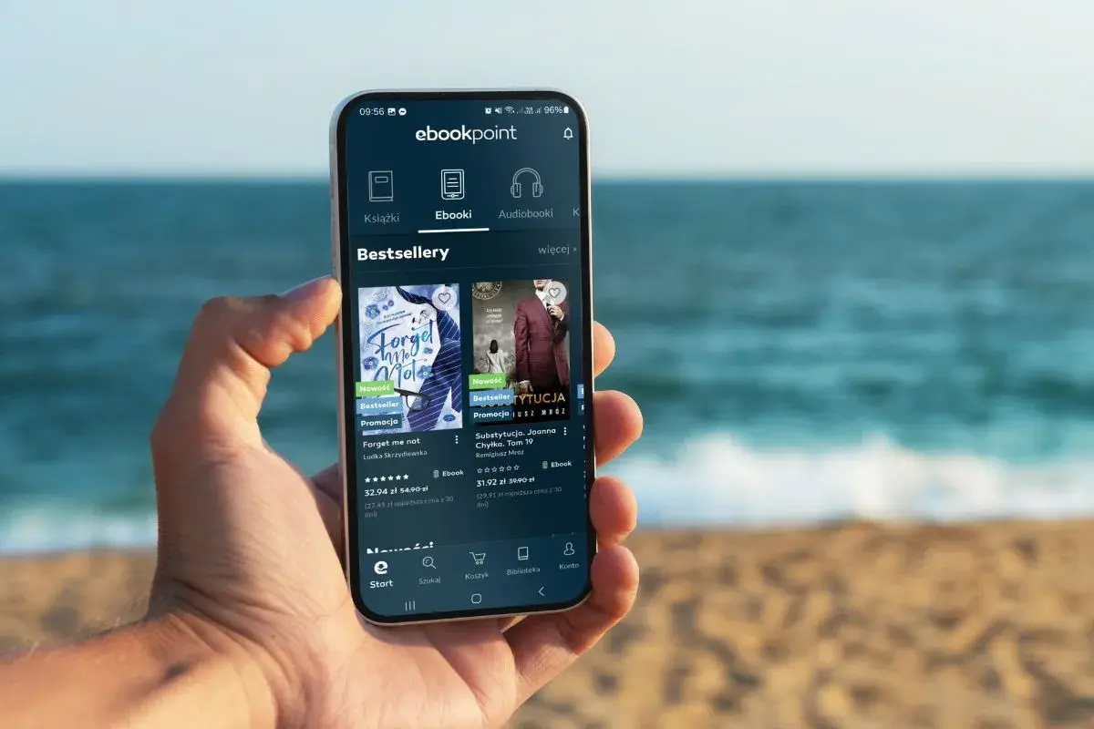 Na smartfonie w dłoni wyświetla się aplikacja ebookpoint z bestsellerami. Idealny poradnik przetrwania na wakacjach, by nie nudzić się na plaży.