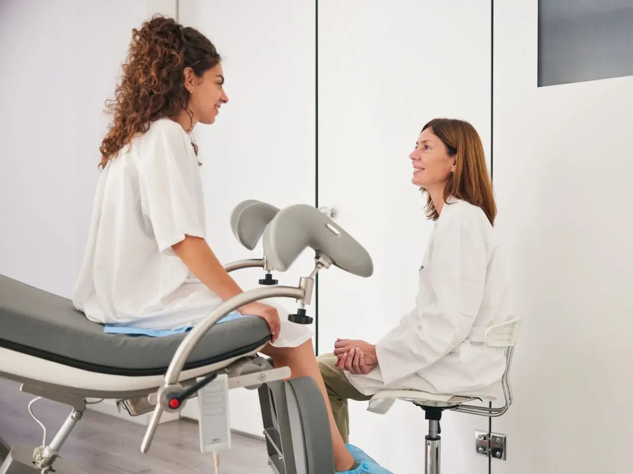 Frauenärztin im Gespräch mit Patientin auf dem gynäkologischen Stuhl. Auch während der Periode ist ein Besuch beim Frauenarzt möglich und wichtig.