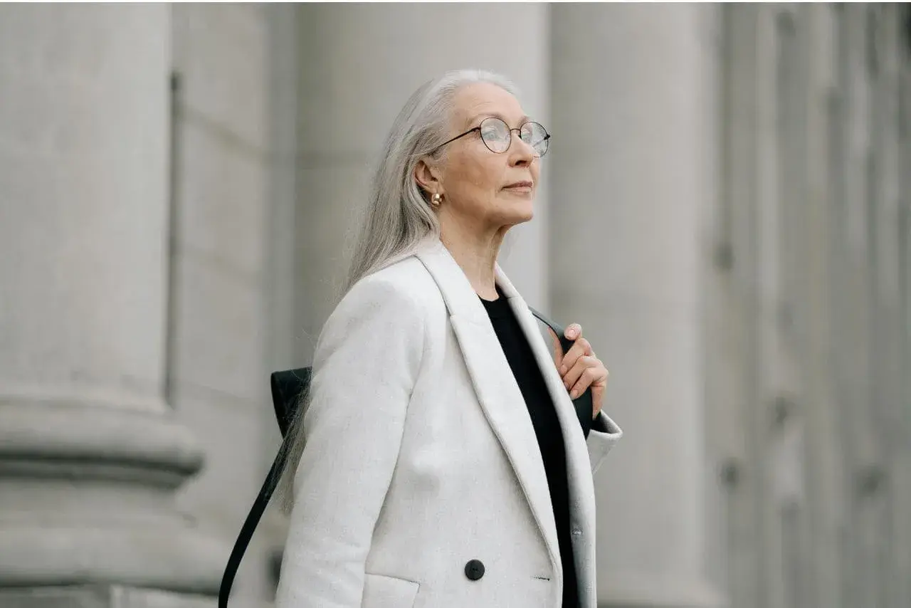 Donna con lunghi capelli grigi, occhiali e un cappotto bianco elegante, con uno sfondo architettonico in tono grigio.