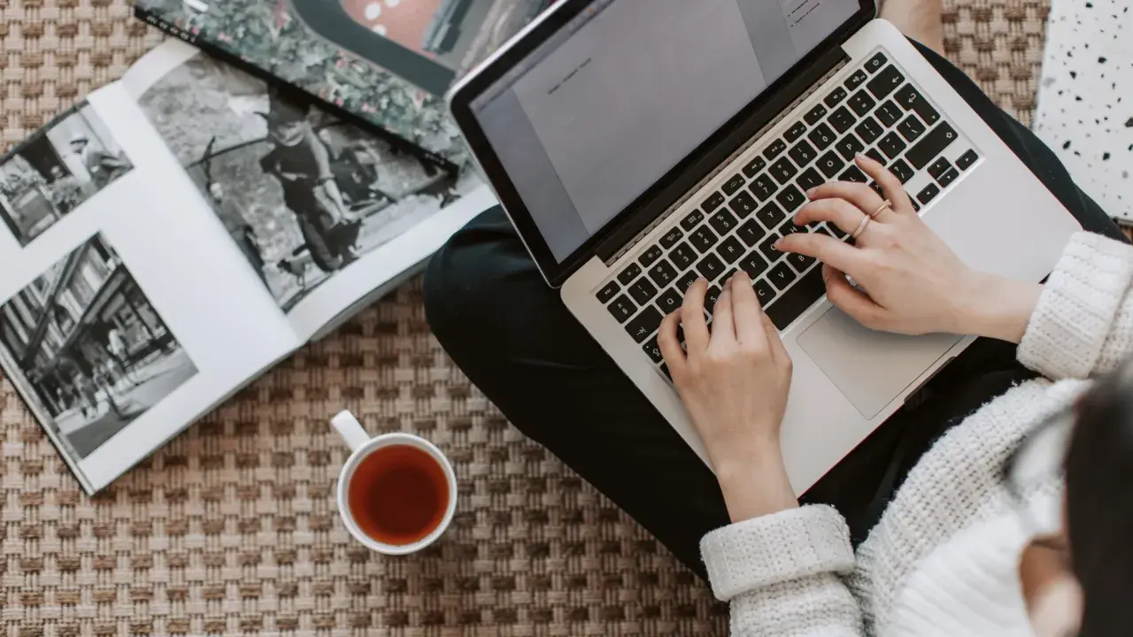Kobieta pisze na laptopie, obok leży otwarta księga i filiżanka herbaty. Zastanawia się, ile kosztuje artykuł sponsorowany.