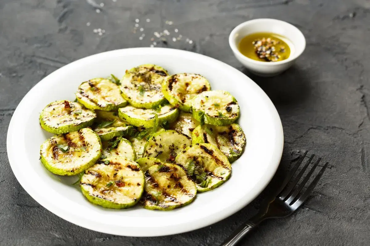 Zucchine alla scapece non fritte, grigliate e condite con olio e spezie, servite su un piatto bianco con una ciotolina di condimento.