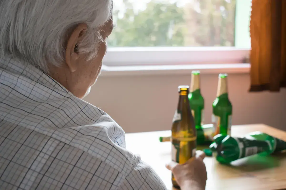 Starszy mężczyzna zmagający się z alkoholizmem, siedzi przy stole z kilkoma pustymi butelkami po piwie.