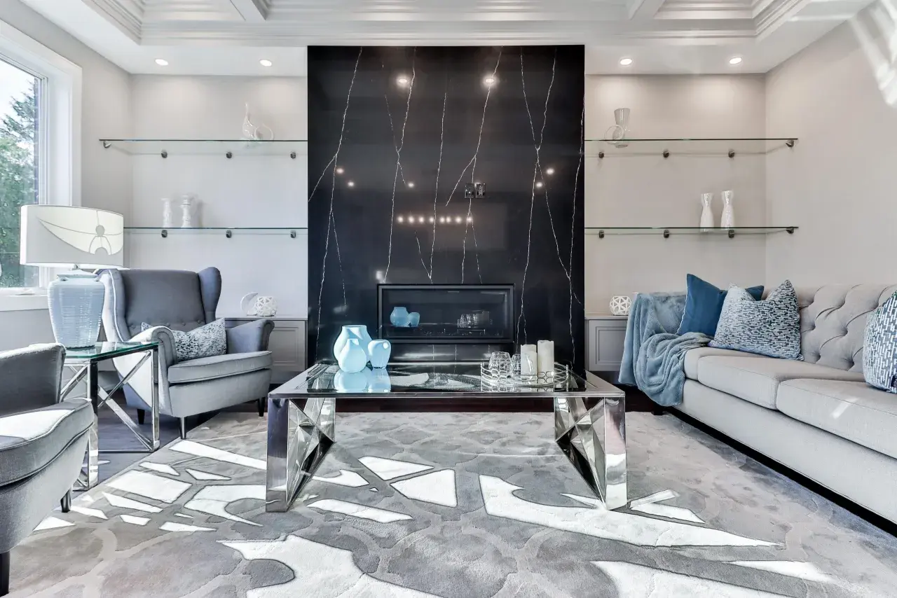 Elegancki salon z kominkiem obłożonym czarnym marmurem i **ścianami w stylu glamour**. Szare fotele, sofa i szklane półki tworzą luksusową atmosferę.