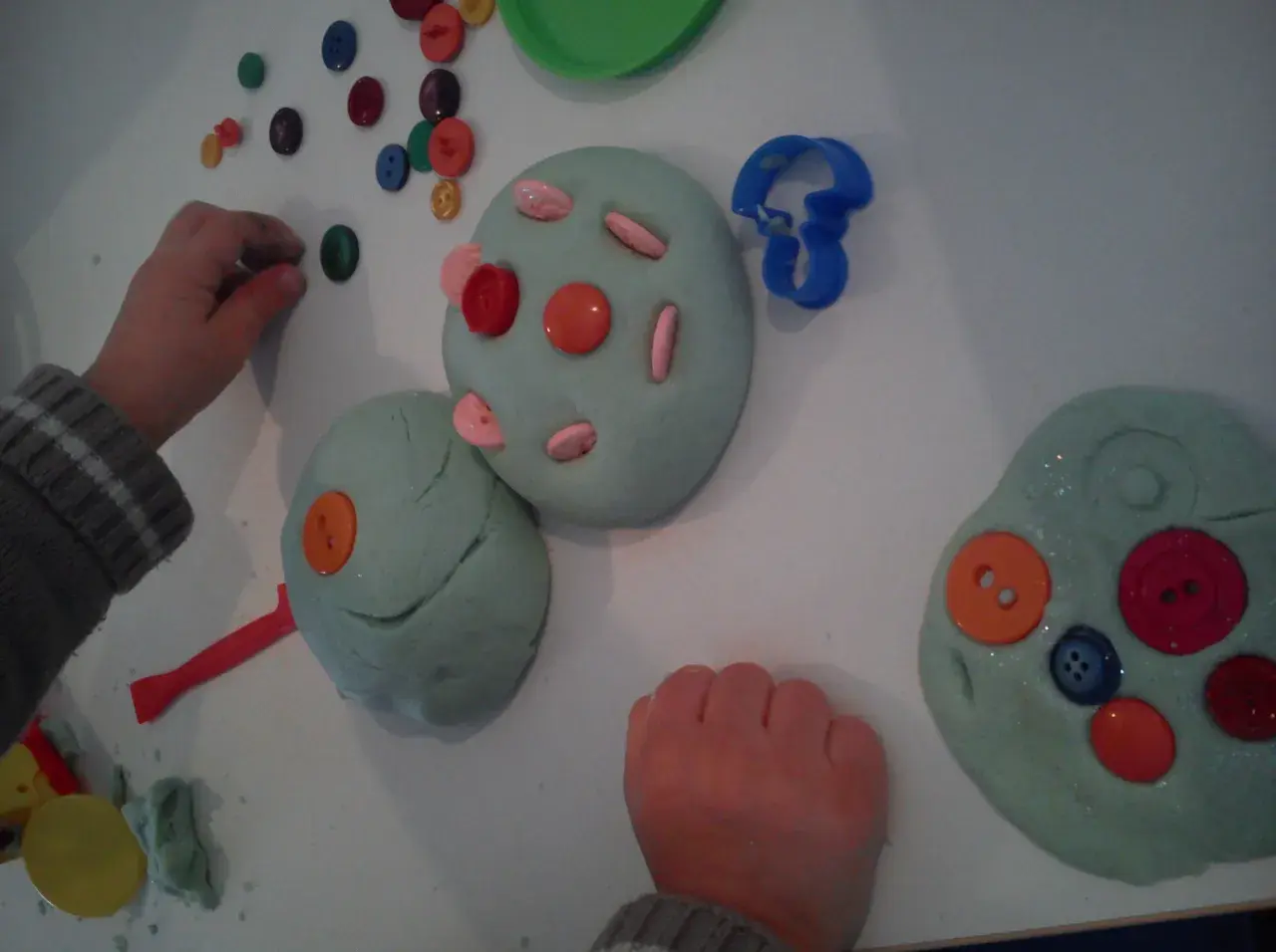 Manos infantiles jugando con plastilina casera, decorada con botones de colores y cortadores.