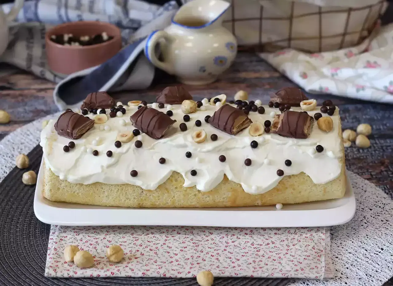 Puszysty biszkopt z kremem, udekorowany kawałkami batoników Kinder Bueno White, orzechami i czekoladowymi kuleczkami.