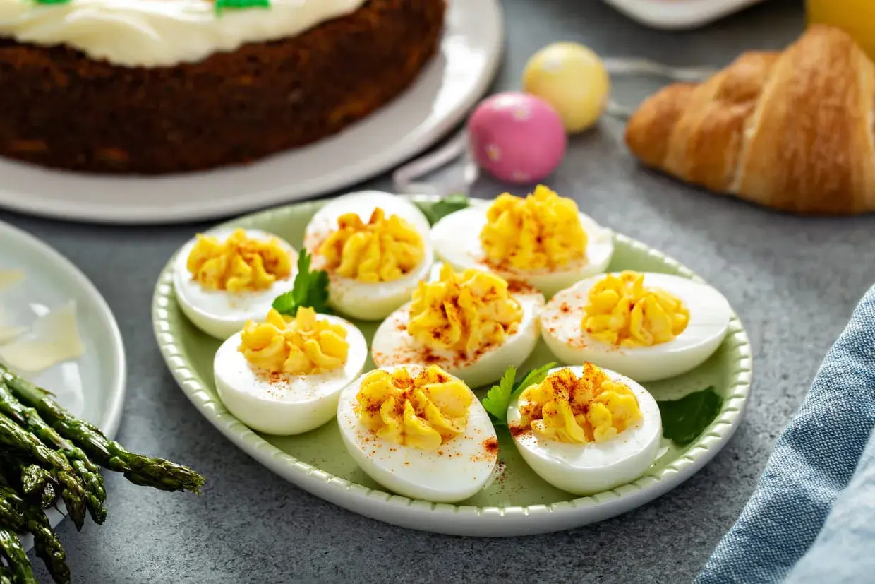 Une assiette d'œufs mimosa, une entrée traditionnelle de Pâques, avec un gâteau aux carottes et des œufs décorés en arrière-plan.
