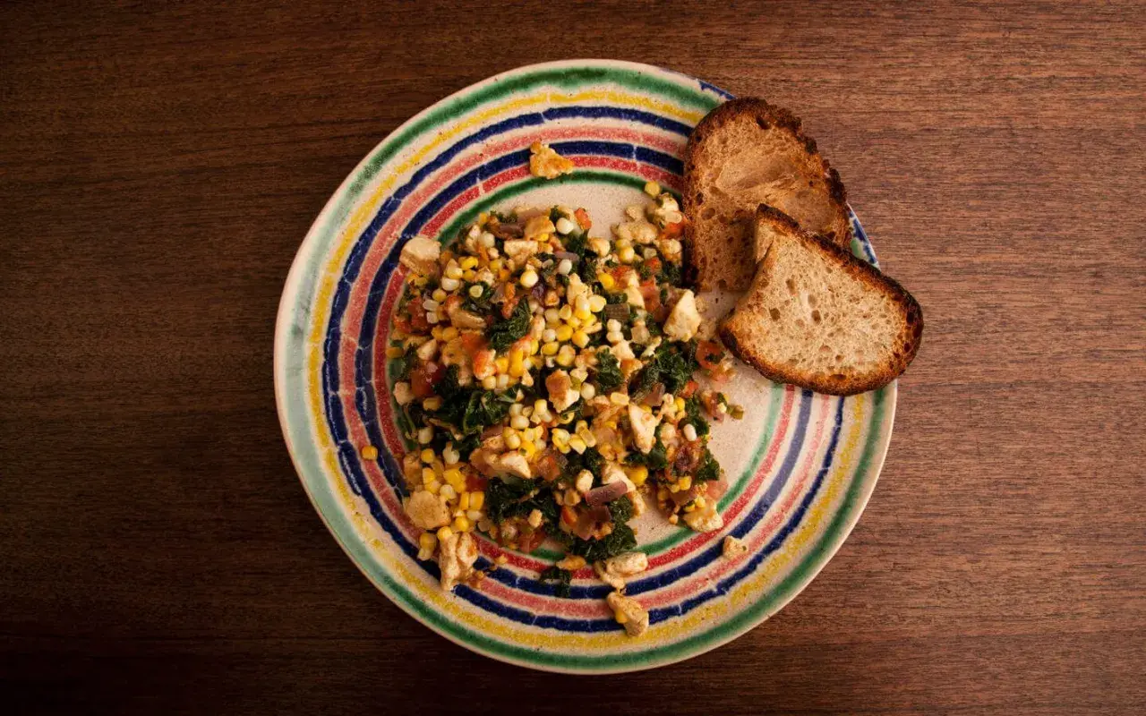 Revuelto de tofu con maíz y espinacas, servido con pan tostado en un plato colorido.