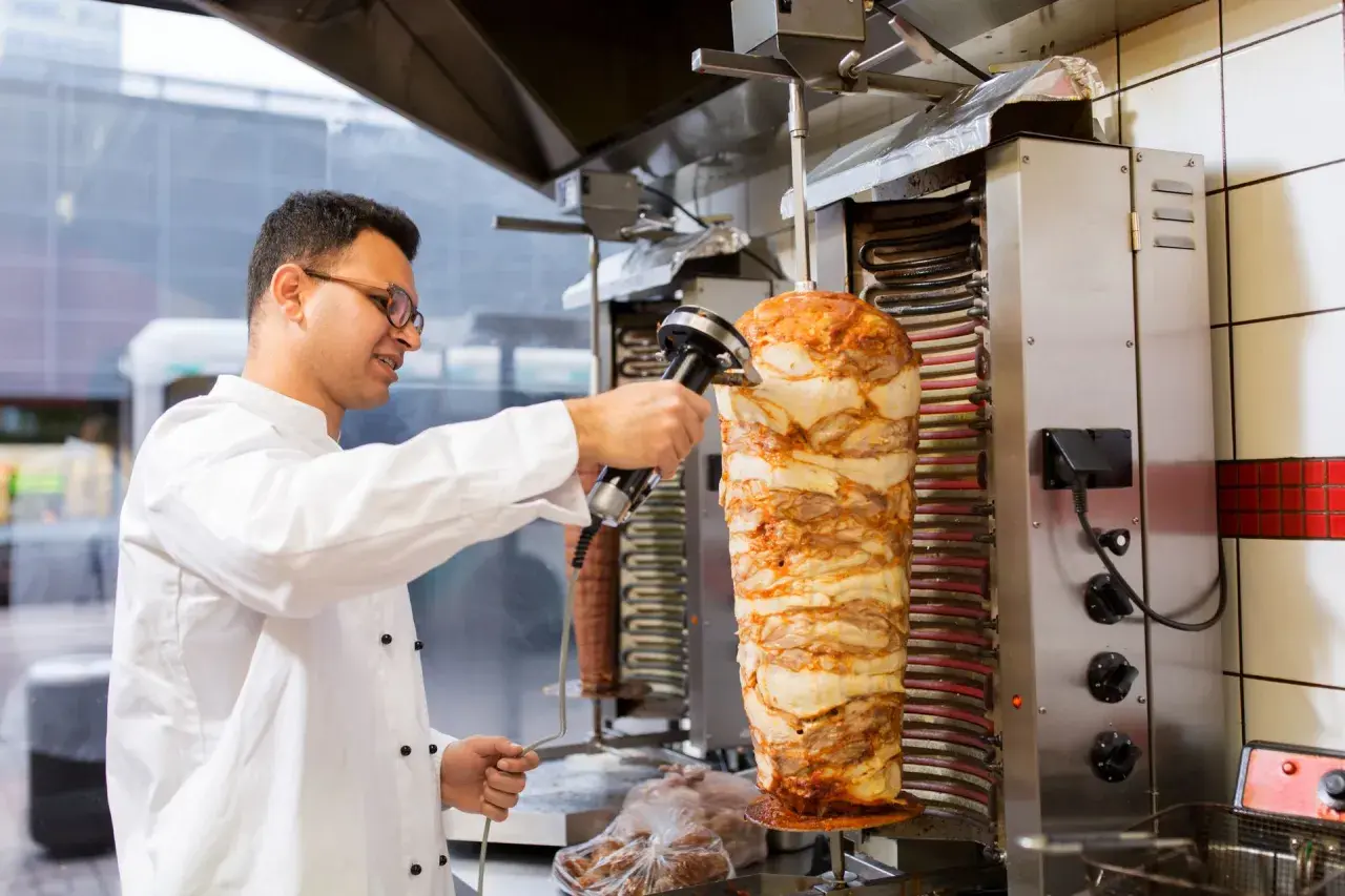 Kucharz w białej kurtce kroi mięso z pionowego rożna na kebab.
