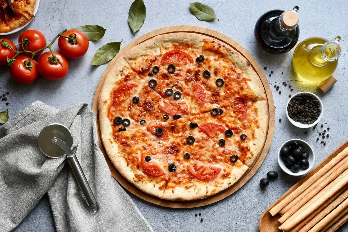Pyszna pizza bez glutenu z pomidorami i oliwkami, gotowa do pokrojenia. Obok świeże pomidory, oliwa i grissini.