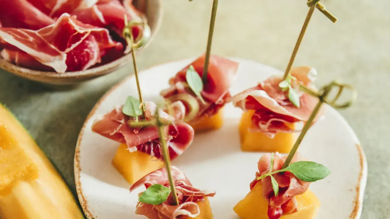 Une présentation originale de melon et jambon, des brochettes savoureuses sur une assiette blanche, avec un melon entier sur le côté.