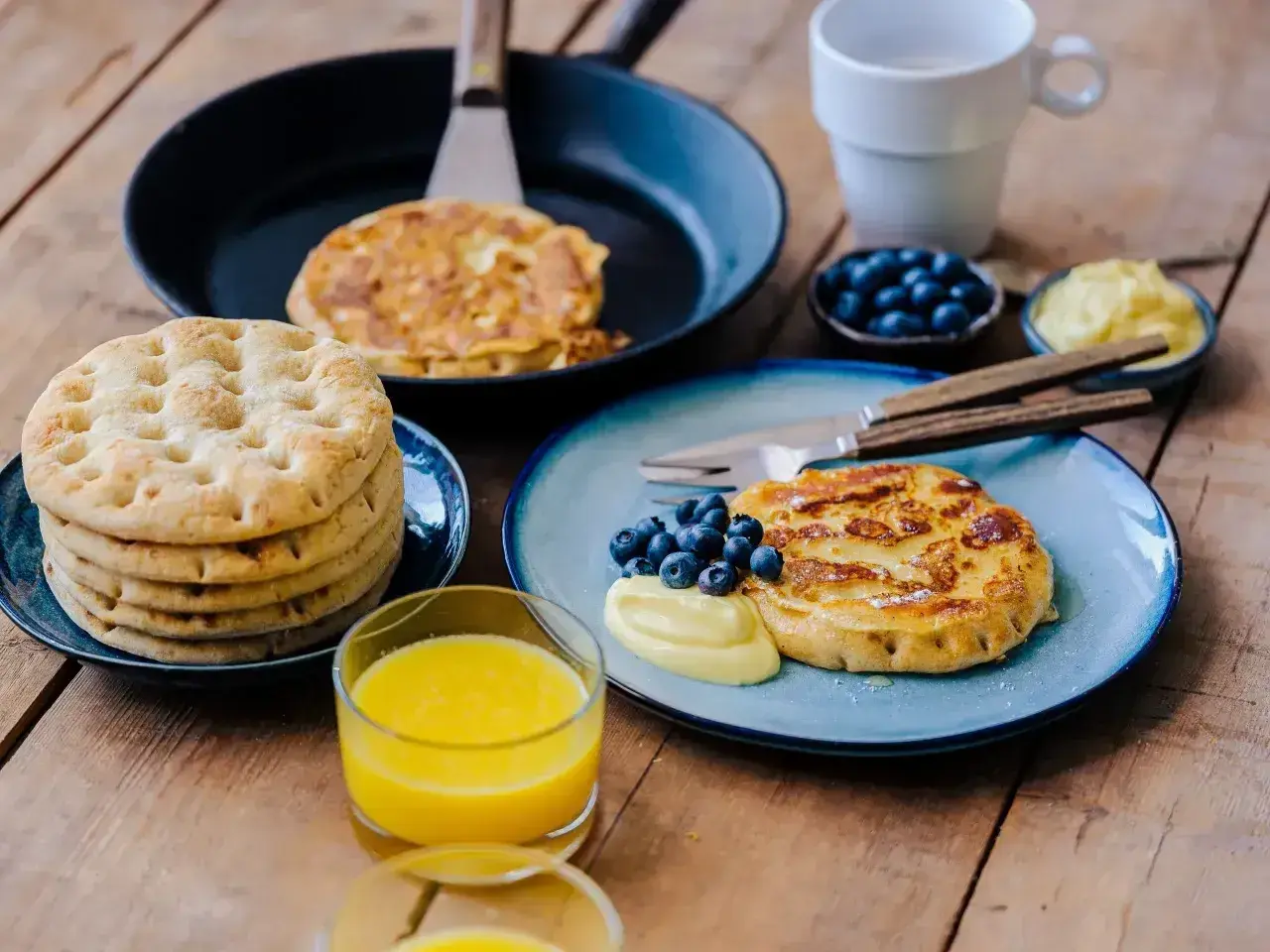 Fattiga riddare med blåbär och vaniljsås på en träyta. En stapel tunna pannkakor och ett glas apelsinjuice.