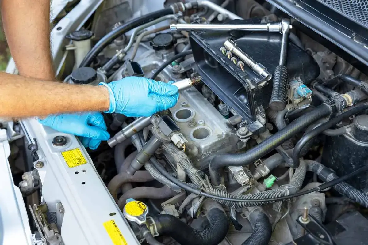 Mano con guante azul sosteniendo una bujía, lista para el cambio de bujías en el motor de un coche. Herramientas a la vista.