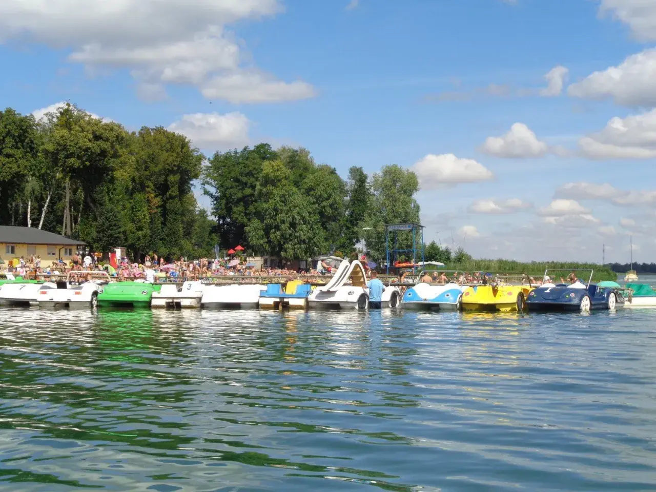 Słoneczny dzień nad jeziorem w Powidzu. Rząd kolorowych rowerów wodnych czeka na plażowiczów.