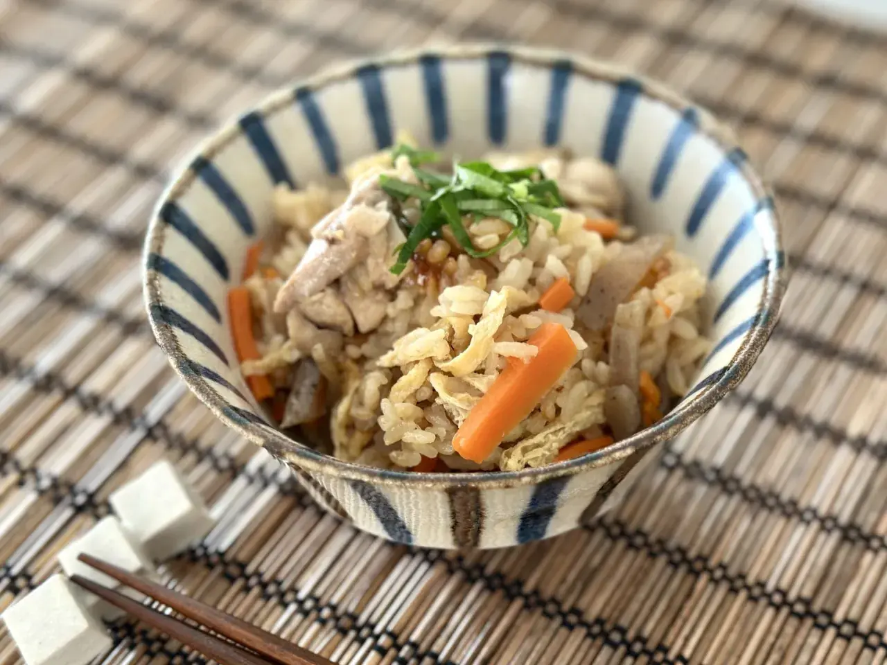 A delicious takikomi gohan recipe served in a striped bowl, featuring rice, chicken, carrots, and green onions.