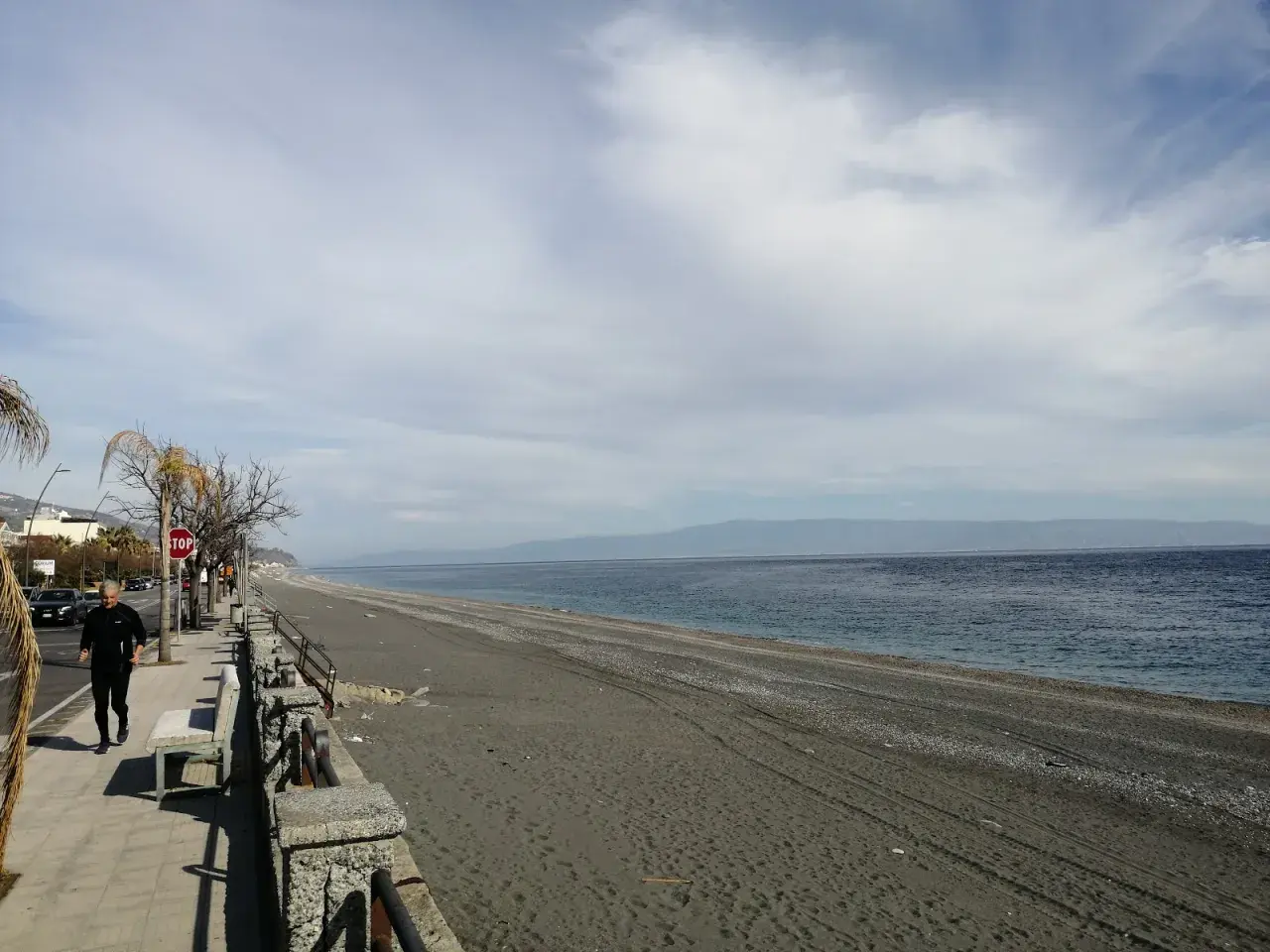 Un uomo corre sul lungomare di Roccafiorita, la spiaggia scura si estende verso l'orizzonte sotto un cielo nuvoloso.