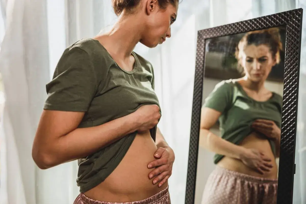 Mujer se mira al espejo, tocándose el abdomen con preocupación por la hinchazón después de comer.