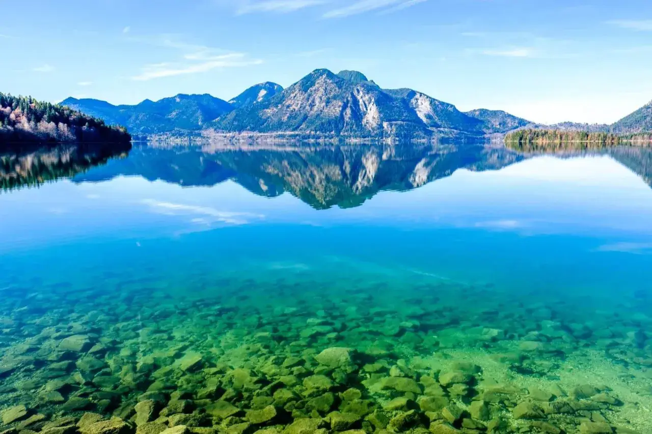 Kristallklares Wasser mit Steinen am Grund, Berge spiegeln sich im See. Perfekt für einen Badeurlaub in Bayern.