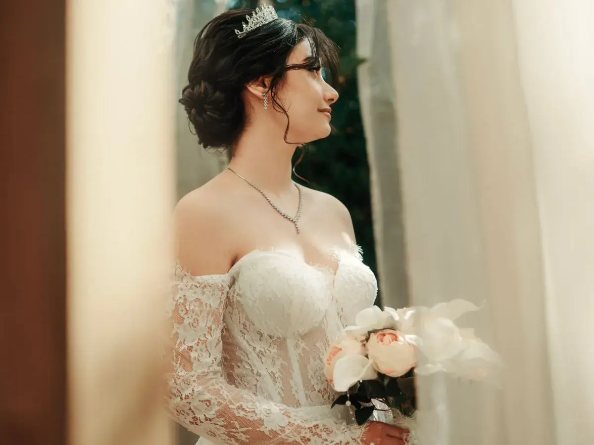 Eleganckie, najmodniejsze upięcia fryzury na wesele. Panna młoda w tiarze i sukni z koronki trzyma bukiet.
