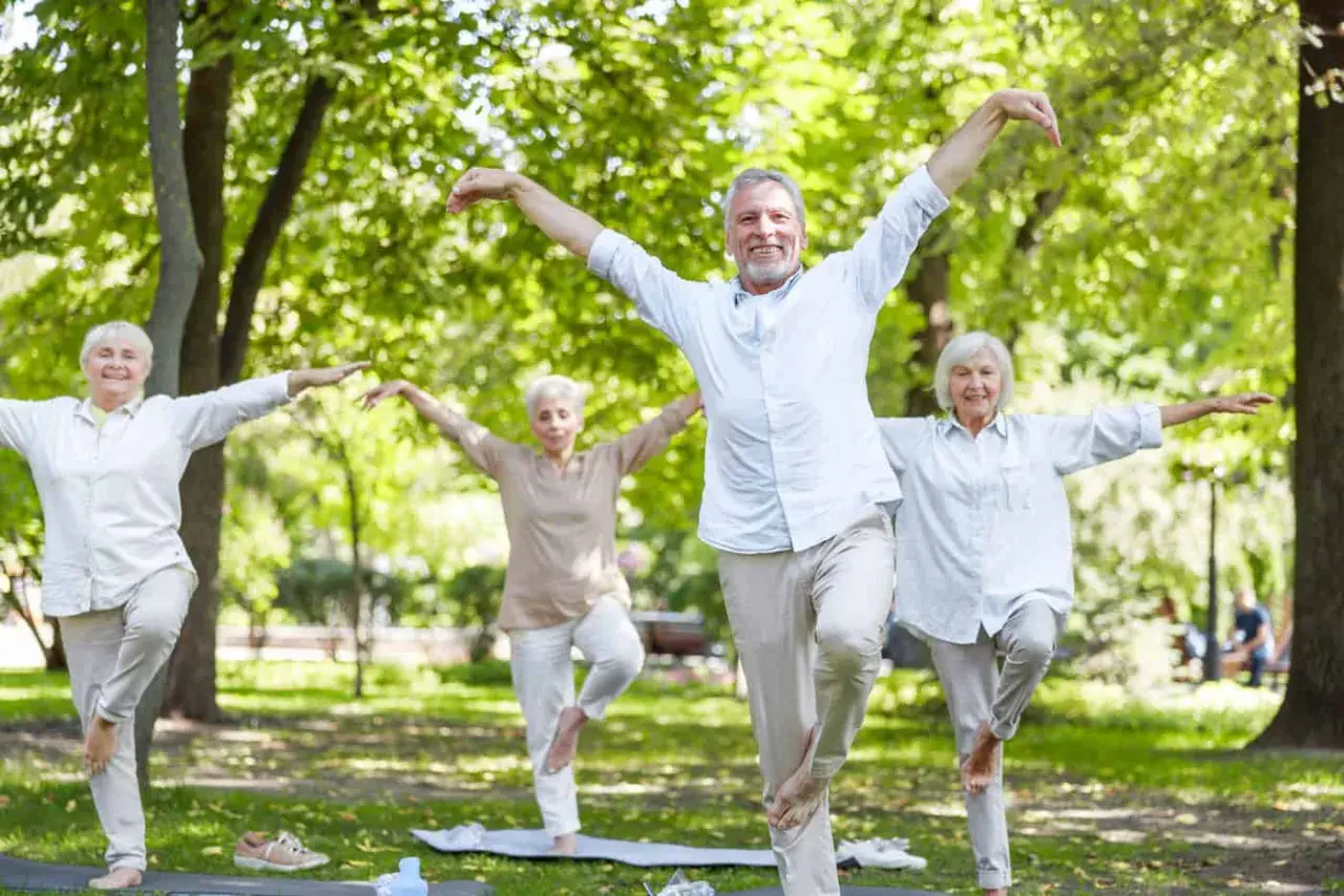 Senioren sport im Park: Vier ältere Menschen balancieren auf einem Bein, Arme ausgebreitet, in einer Yoga-ähnlichen Pose.